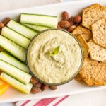 Almond pesto dip in a bowl on a tray surrounded by almonds, crackers and cucumber slices.