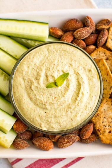 Almond pesto dip in a bowl on a tray surrounded by almonds, crackers and cucumber slices.