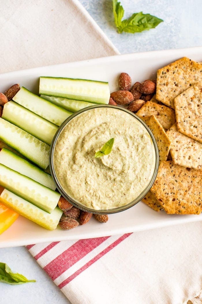 Almond pesto dip in a bowl on a tray surrounded by almonds, crackers and cucumber slices.