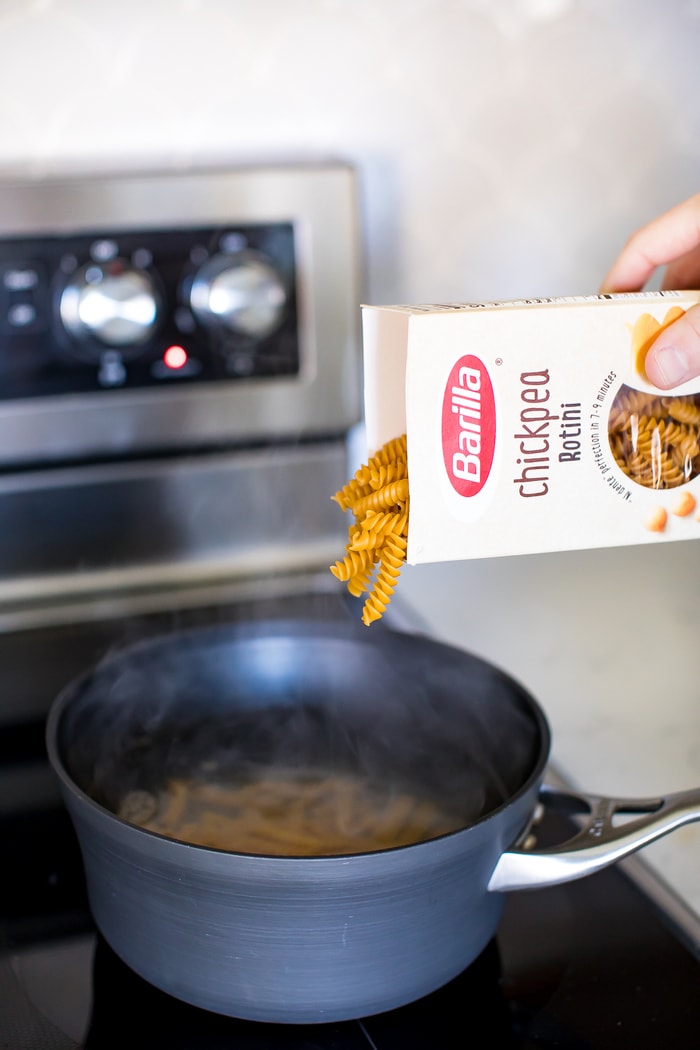 Hand pouring a box of Barilla chickpea rotini pasta into a boiling pot of water for protein mac and cheese.