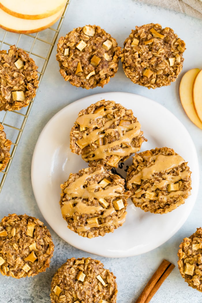 Apple baked oatmeal cups on a white plate with nut butter drizzle, surrounded by more baked apple oatmeal cups, apple slices, and a cinnamon stick.