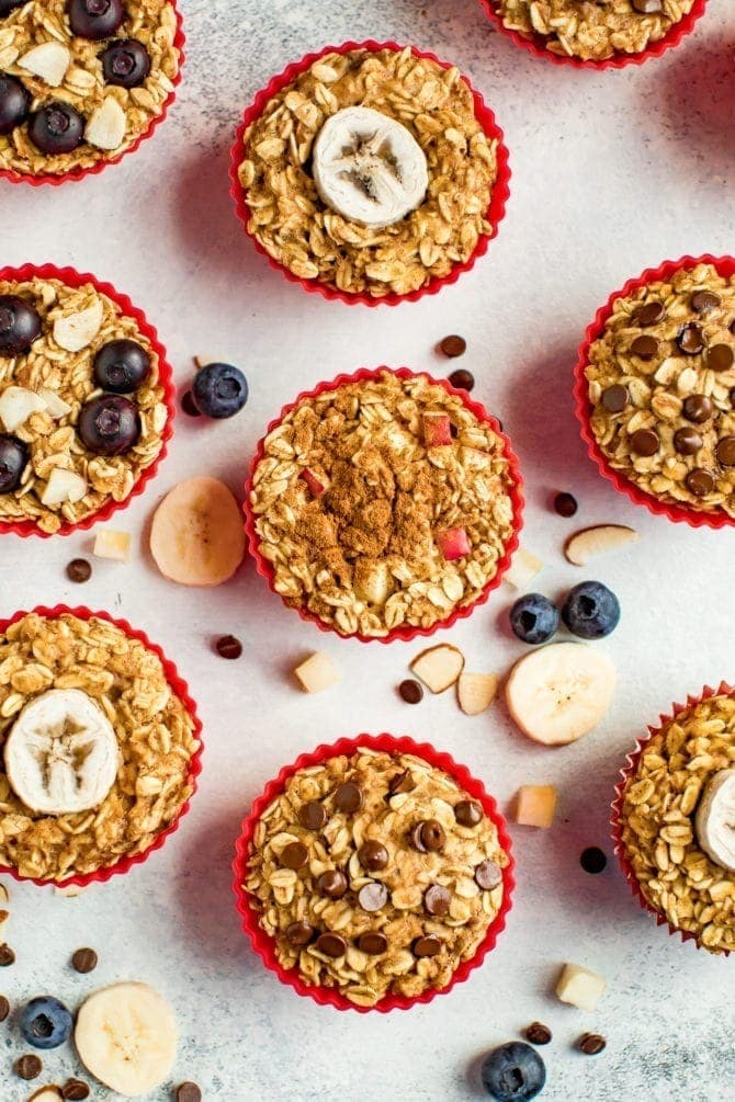 Muffin liners with baked oatmeal cups 4 ways. ingredients are surrounding them on the table like chocolate chips, blueberries, and apple chunks.