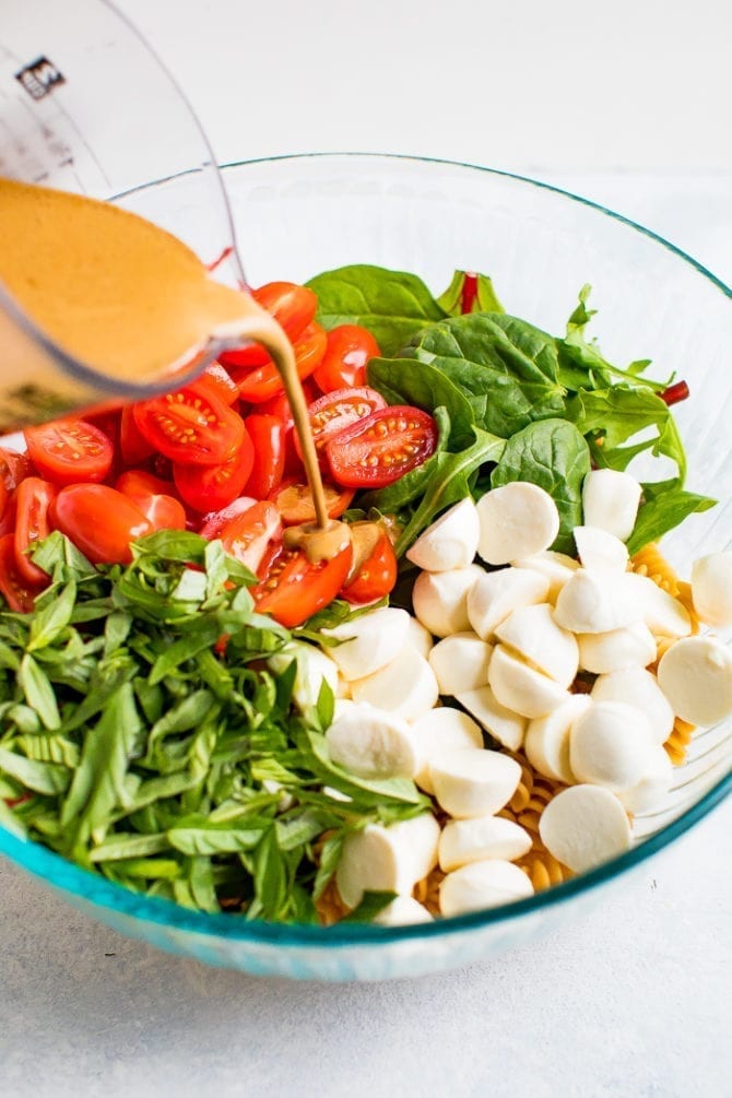 Serving bowl of gluten free caprese pasta salad with tomatoes, mozzarella, basil and a balsamic vinaigrette being poured over the top.