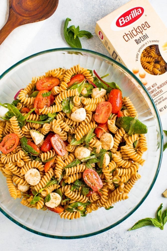 Serving bowl of gluten free caprese pasta salad with tomatoes, mozzarella, basil and a balsamic vinaigrette. Box of Barilla chickpea pasta.