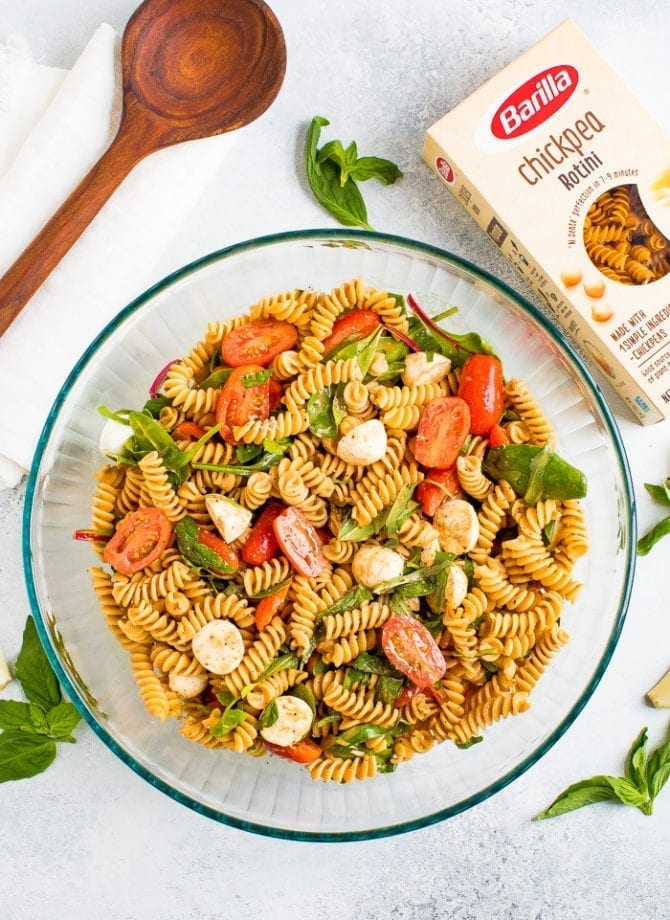 Serving bowl of gluten free caprese pasta salad with tomatoes, mozzarella, basil and a balsamic vinaigrette. Box of Barilla chickpea pasta is to the side.