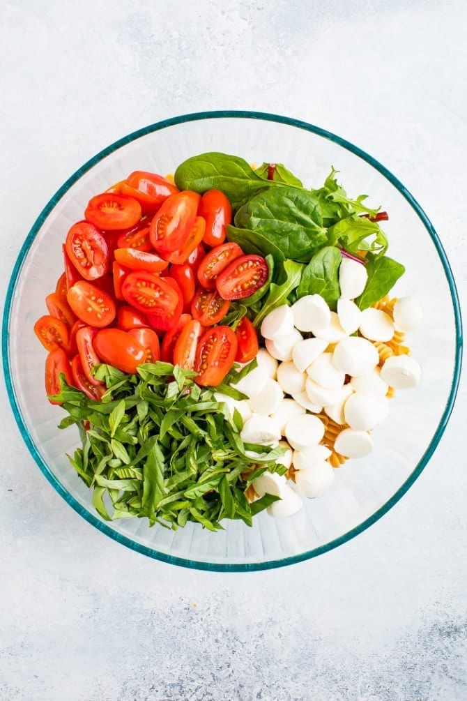 Ingredients in a bowl for gluten free caprese pasta salad including chickpea pasta, cherry tomatoes, basil, fresh mozzarella and greens.