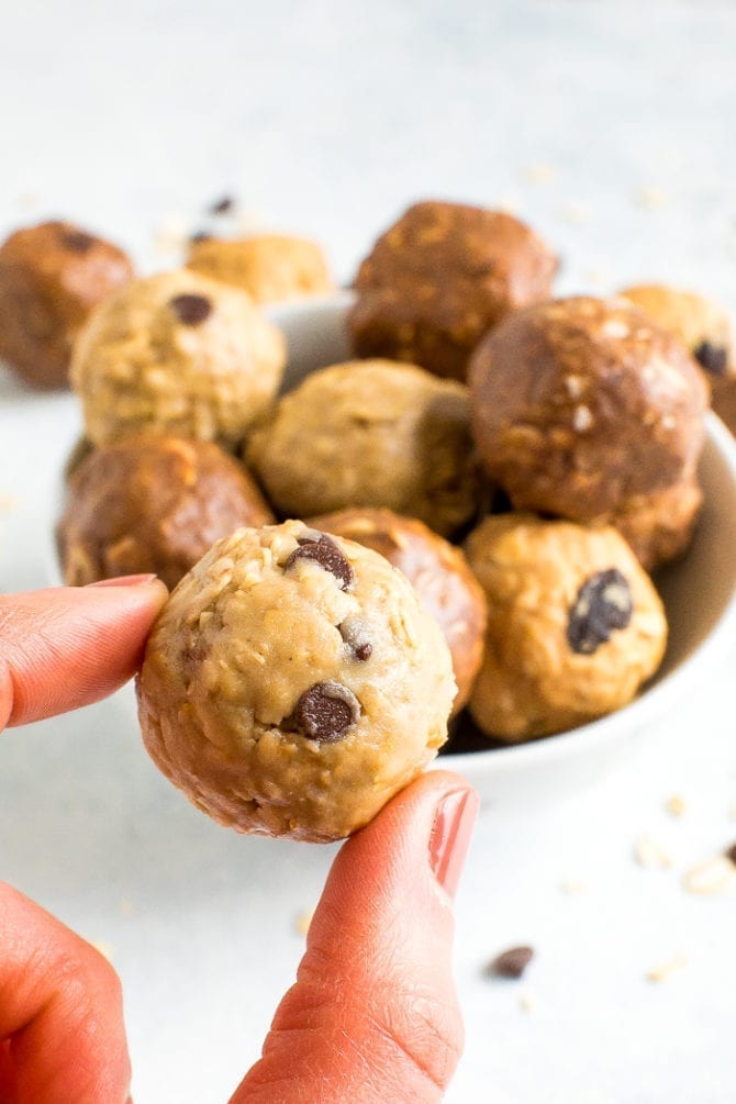 Hand holding a chocolate chip peanut butter protein ball. Behind is a bowl of a variety of protein balls.