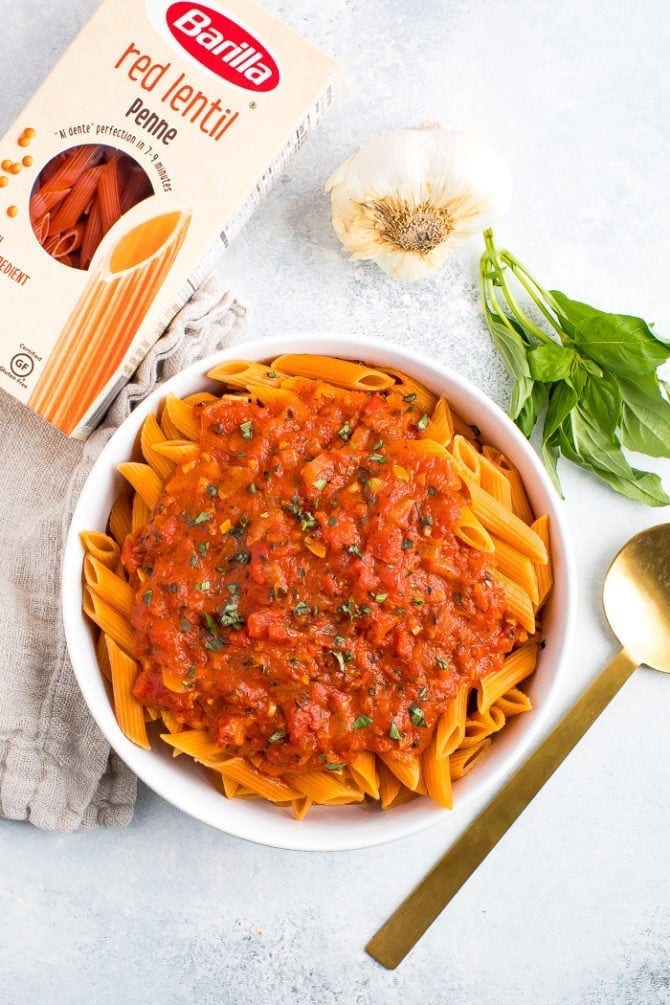 Serving bowl of healthy, vegan and gluten free penne alla vodka next to a box of Barilla red lentil penne, garlic and fresh basil.