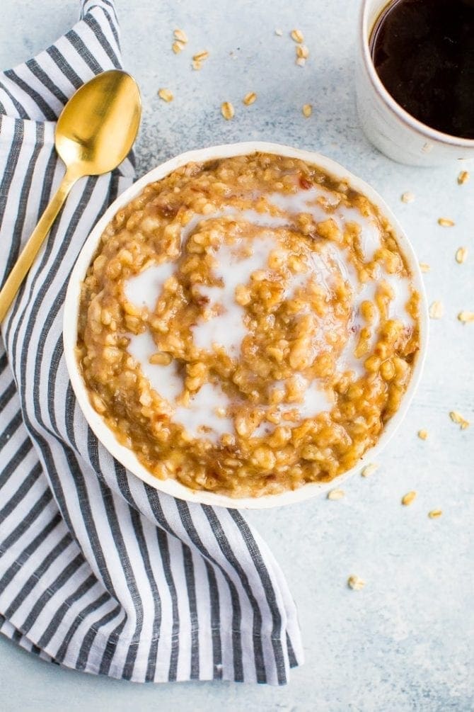 Bowl of salted caramel oatmeal swirled with date caramel and almond milk. A gold spoon, dish cloth, and a mug of coffee are beside the bowl.