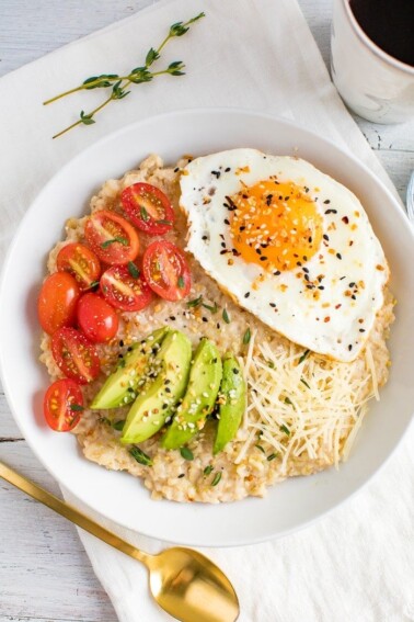 Savory oatmeal topped with avocado, tomatoes, herbs, a fried egg, parmesan, and everything bagel seasoning. Gold spoon and sprigs of thyme on the side.