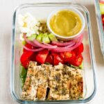 Glass meal prep container with grilled chicken tomatoes, pickled onions, cucumber, quinoa, greens, feta and a lemon vinaigrette.
