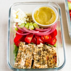 Glass meal prep container with grilled chicken tomatoes, pickled onions, cucumber, quinoa, greens, feta and a lemon vinaigrette.