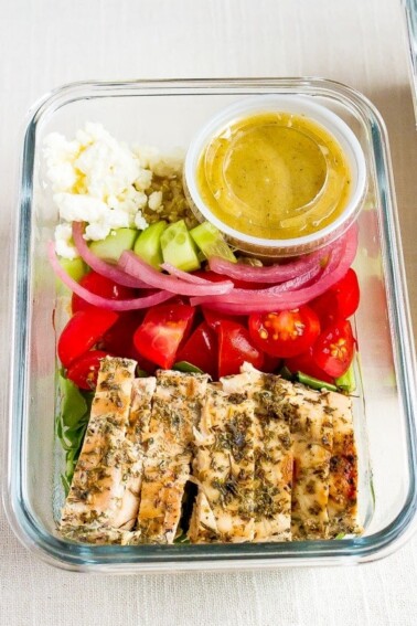 Glass meal prep container with grilled chicken tomatoes, pickled onions, cucumber, quinoa, greens, feta and a lemon vinaigrette.