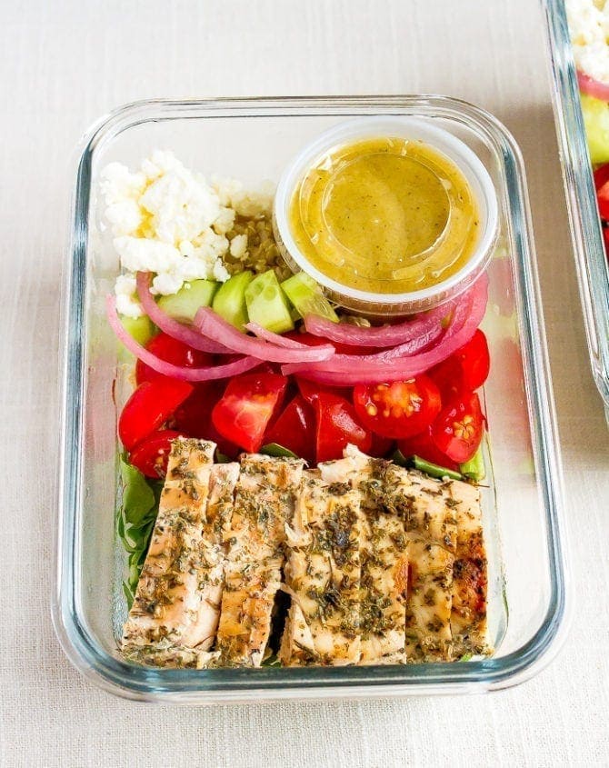 Glass meal prep container with grilled chicken tomatoes, pickled onions, cucumber, quinoa, greens, feta and a lemon vinaigrette.