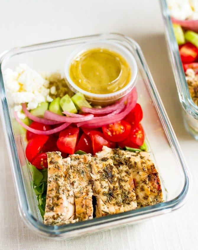 Glass meal prep container with grilled chicken tomatoes, pickled onions, cucumber, quinoa, greens, feta and a lemon vinaigrette.