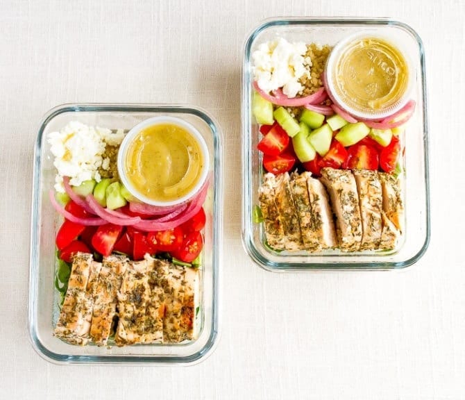 Two grilled chicken meal prep bowls with tomatoes, onion, cucumber, feta, quinoa, greens, and a vinaigrette.