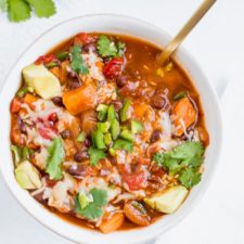 Slow cooker sweet potato and black bean vegetarian chili in a bowl topped with cilantro, cheese and jalapeño with a spoon on a white table.