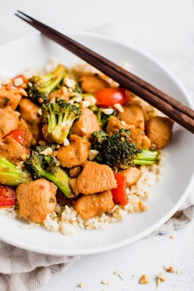 Kung pao chicken over rice with vegetables and peanuts on a plate with chopsticks.