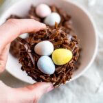 A hand holding a chocolate coconut flake Easter nest treat with Cadbury eggs in the nest.