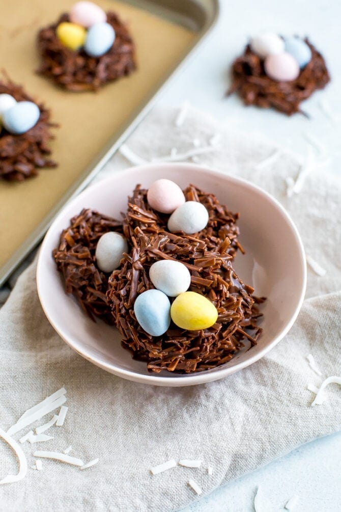 The chocolate coconut nests with Cadbury eggs in a white bowl, one nest on the table and two on parchment on a baking sheet.