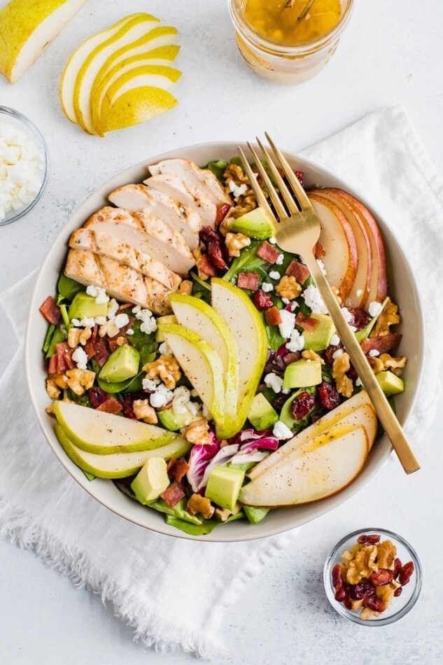Salad topped with pears, grilled chicken, turkey bacon, cranberries, goat cheese, walnuts, and avocado with a fork. Dressing, fork, cheese in a bowl, slices of pear and walnuts around salad bowl.