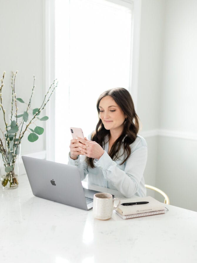 Woman sitting at a kitchen counter, on her phone. Her laptop, coffee mug, and notebook are on the counter.