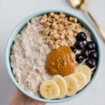Greek yogurt oatmeal with bananas, blueberries, granola and peanut butter on top. with a gold spoon on the side.