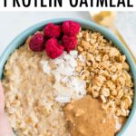 Hand holding a bowl of oatmeal topped with raspberries, granola, peanut butter and coconut.