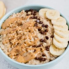 Oatmeal topped with nut butter drizzle, chocolate chips, and sliced banana in a blue bowl.