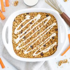 Carrot cake baked oatmeal in a baking dish drizzled with cream cheese frosting, surrounded by a whisk, catto sticks, and walnuts.