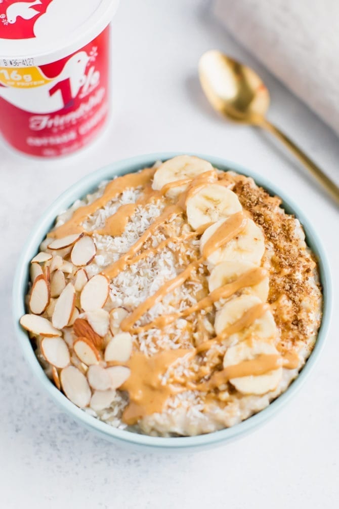 Bowl of cottage cheese protein oatmeal topped with sliced almonds, coconut flakes, banana, flax seeds and peanut butter. Cottage cheese container and spoon are in the background.