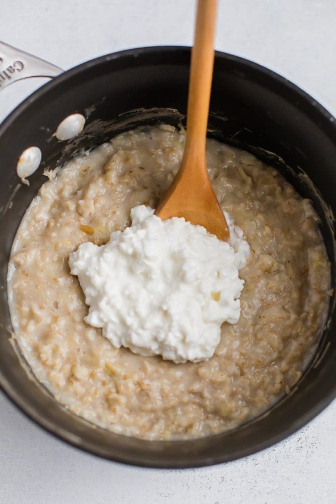 Pot of oatmeal with cottage cheese being stirred in with a wooden spoon.