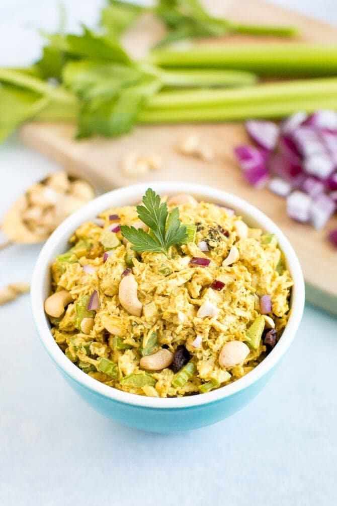 Bowl of curry chicken salad. Cutting board in the background with onion, celery, and cashews.