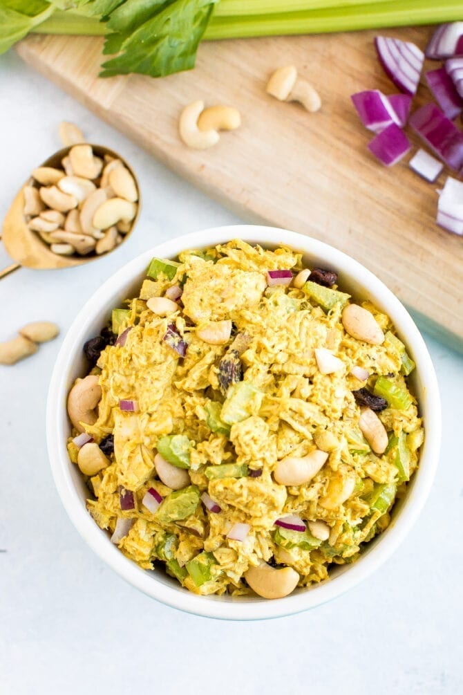 Bowl of curry chicken salad. Cutting board in the background with onion, celery, and cashews.