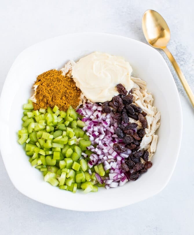 Mixing bowl with a gold spoon beside it. Ingredients for the curry chicken salad are in the bowl: curry powder, mayo, shredded chicken, raisins, chopped onion, chopped celery.