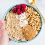 A bowl of egg white oatmeal with granola, freeze dried raspberries, coconut and almond butter in a blue bowl with a spoon.