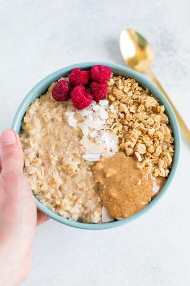 A bowl of egg white oatmeal with granola, freeze dried raspberries, coconut and almond butter in a blue bowl with a spoon.