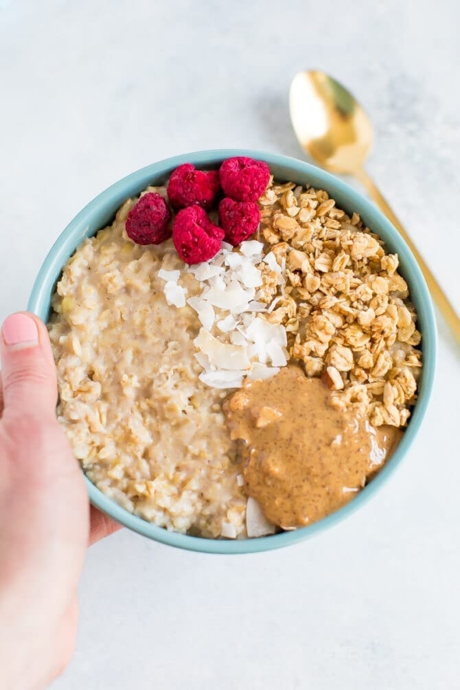 Hand holding a bowl of egg white oatmeal topped with peanut butter, granola, coconut, and freeze dried raspberries.