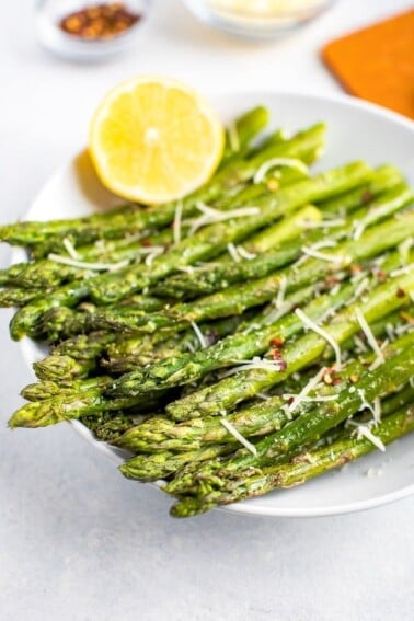 Roasted asparagus on a plate topped with red pepper flakes, parmesan and fresh lemon.