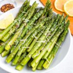 Plate of roasted asparagus topped with parmesan, red pepper flakes, and a lemon wedge on the side. Beside the bowl are smaller bowls filled with red pepper, parmesan, and a cutting board with lemon slices.