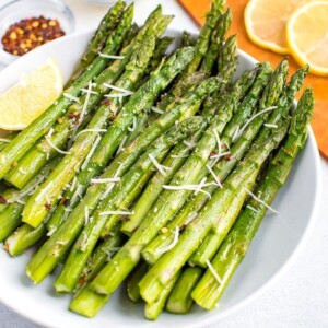 Plate of roasted asparagus topped with parmesan, red pepper flakes, and a lemon wedge on the side. Beside the bowl are smaller bowls filled with red pepper, parmesan, and a cutting board with lemon slices.