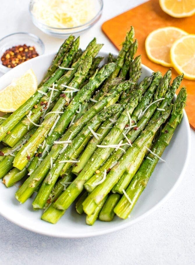 Roasted asparagus on a plate topped with red pepper flakes, parmesan and fresh lemon.