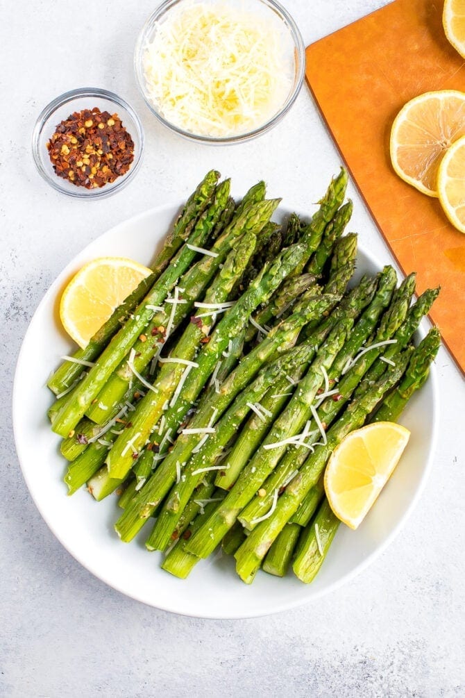 Plate of roasted asparagus topped with parmesan, red pepper flakes, and a lemon wedge on the side. Beside the bowl are smaller bowls filled with red pepper, parmesan, and a cutting board with lemon slices.