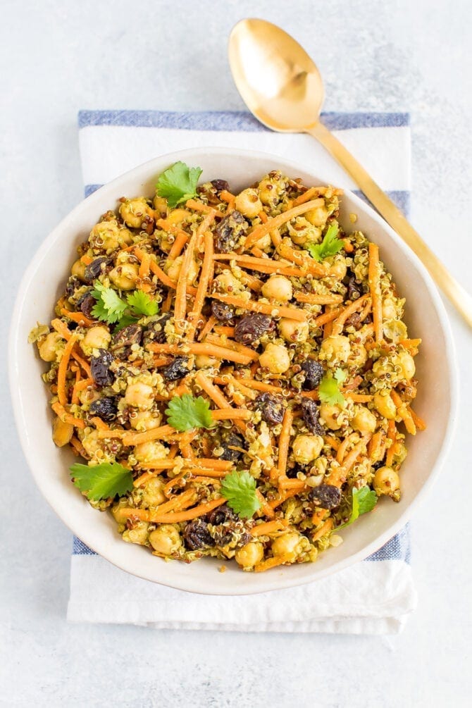 A carrot salad with raisins, quinoa, chickpeas in a bowl with a gold spoon on the right. 