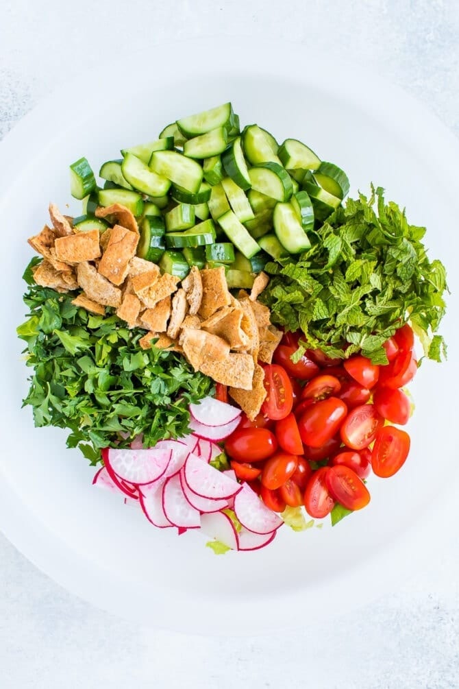 Ingredients for fattoush salad in a bowl before being mixed: parsley, mint, cucumber, crumbled pita chips, tomatoes and radishes.