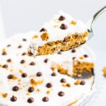 Spatula with a slice of chocolate chip cookie icebox cake, topped with whipped cream and chocolate chips and cookie crumbs. The icebox cake is in the background.