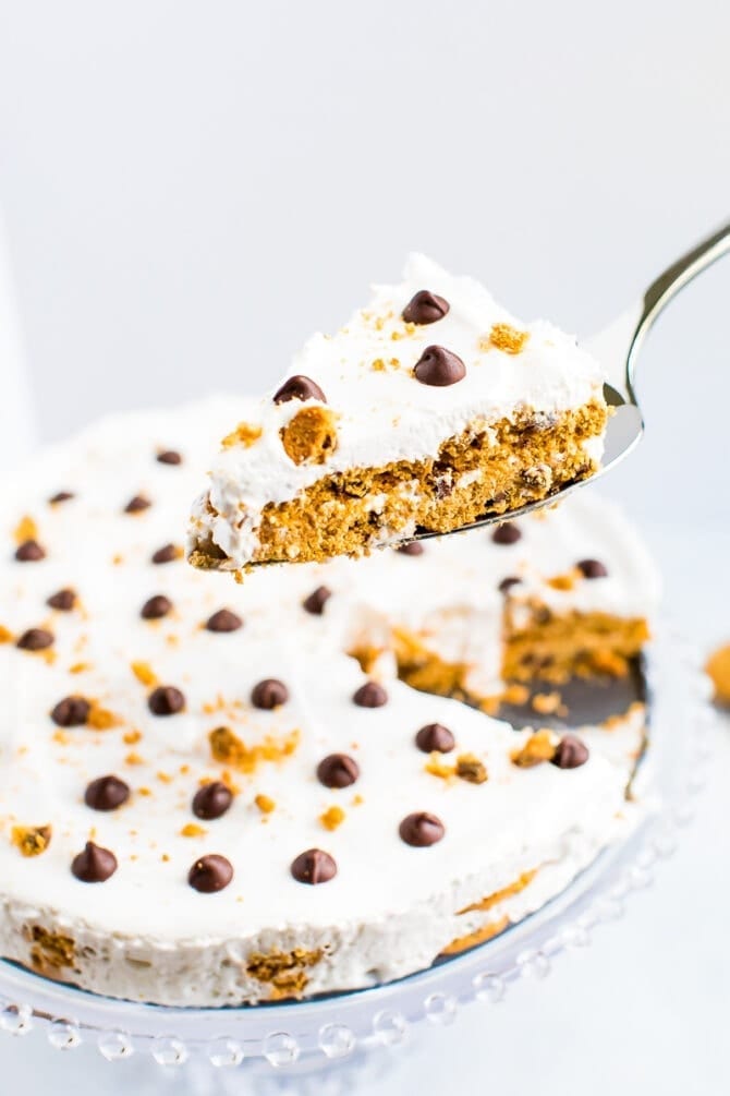 Spatula with a slice of chocolate chip cookie icebox cake, topped with whipped cream and chocolate chips and cookie crumbs. The icebox cake is in the background.