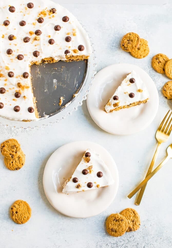 Chocolate chip cookies icebox cake topped with chocolate chips and cookie crumbs. Two slices of the cake are on two plates. Two forks and chocolate chip cookies are on by the plates.