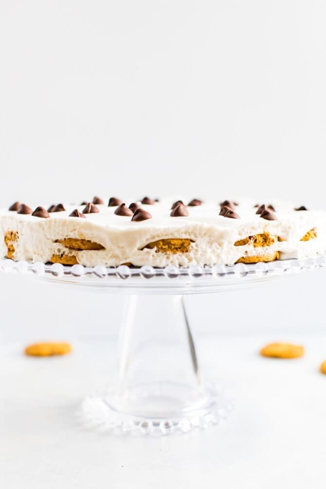 Chocolate chip icebox cake topped with chocolate chips on a glass cake stand. Chocolate chips cookies are on the table.