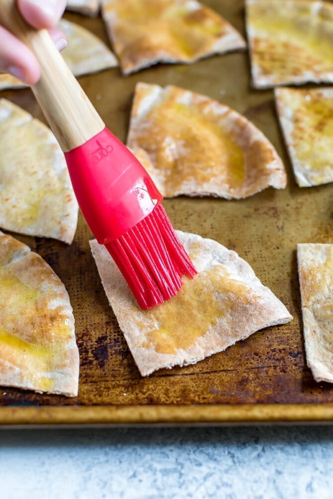 Silicone brush brushing olive oil mixture onto the slices of pita on a baking sheet.